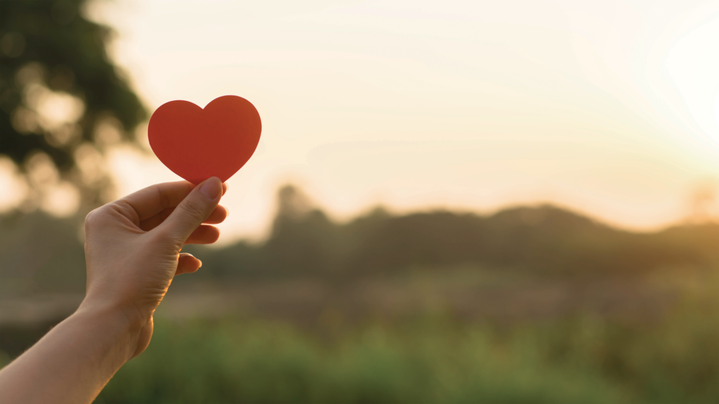 Hand holds a paper heart with a grassy field and sunset in the background