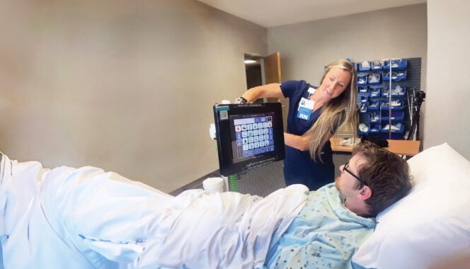 A patient lays in a hospital bed while a nurse shows him how to use an app on a tablet.
