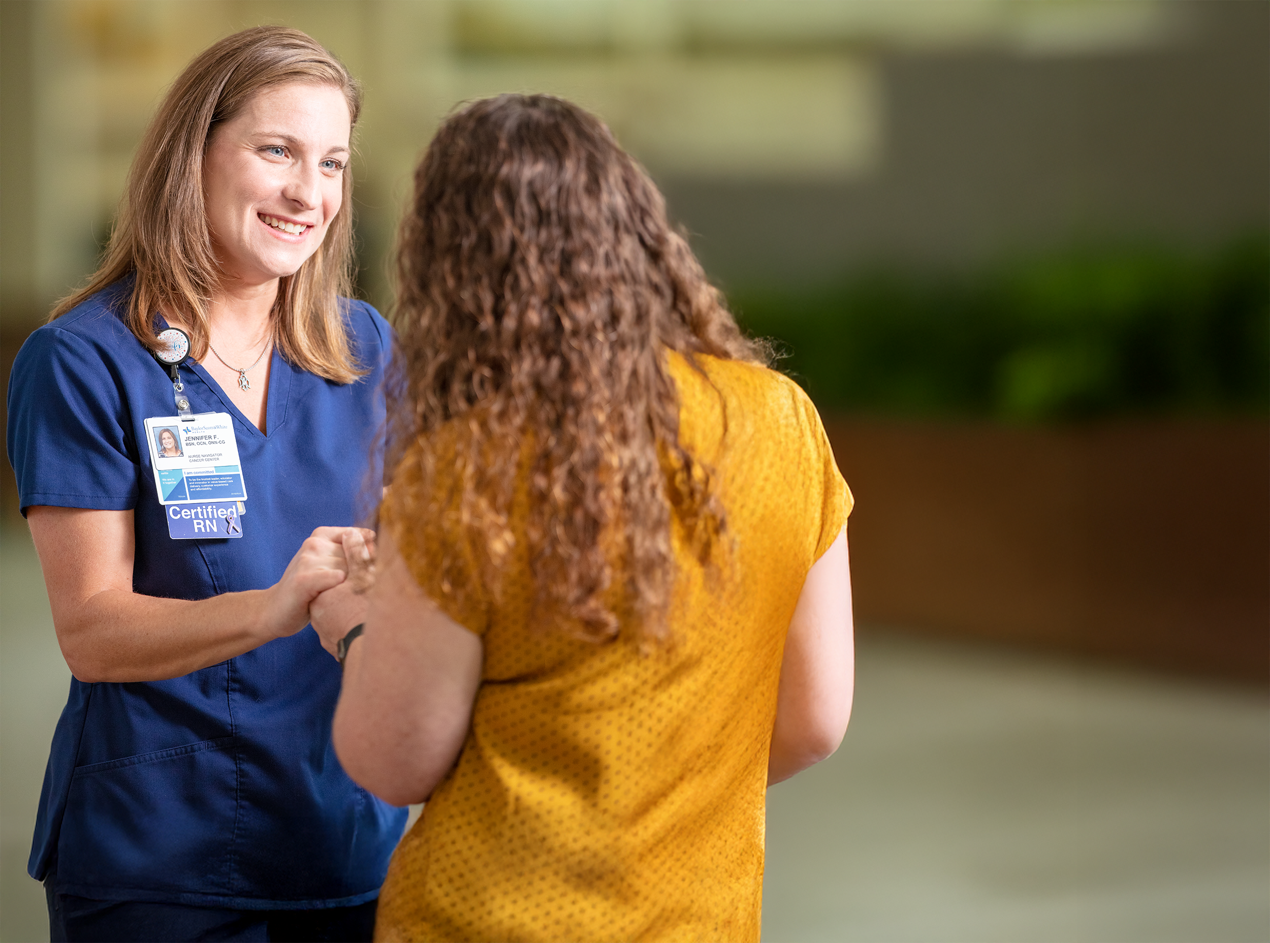 A certified RN in blue scrubs speaks with a person in a yellow shirt inside a healthcare setting.