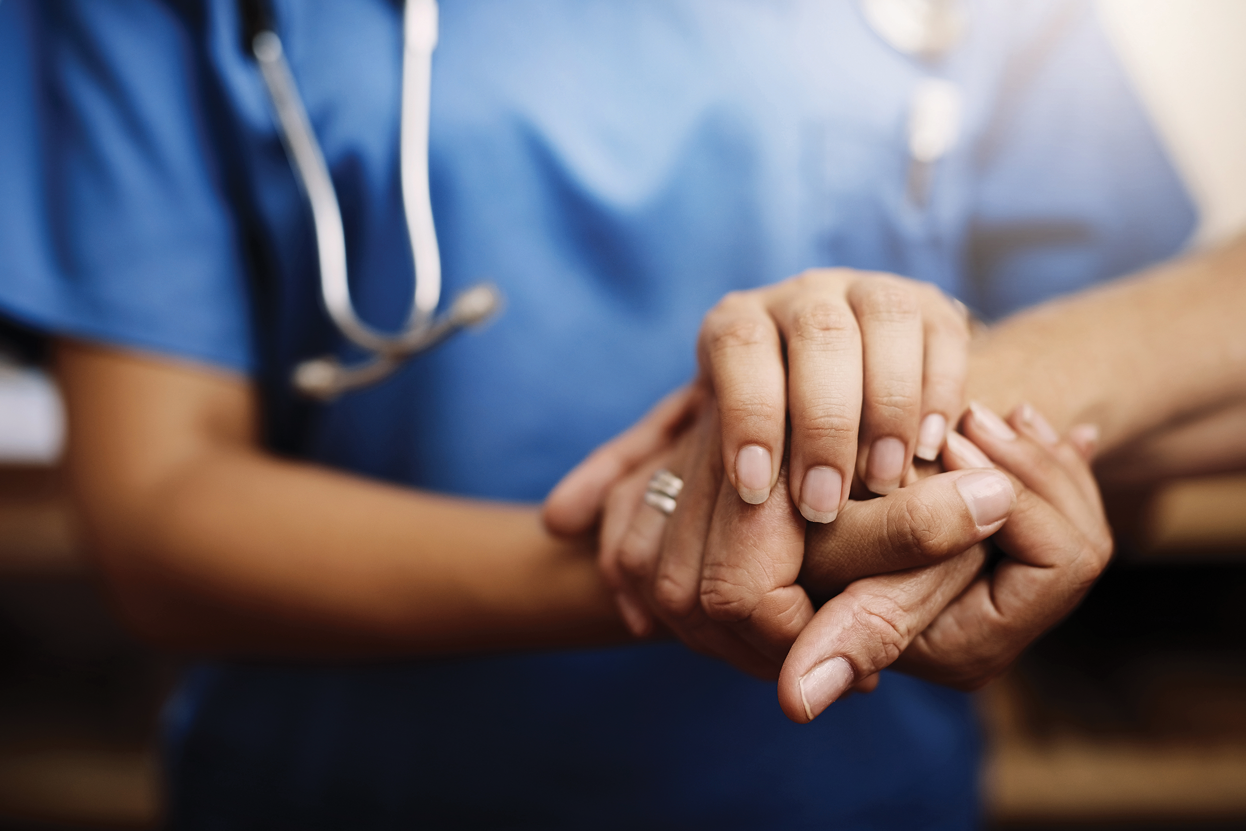 A healthcare worker in blue scrubs gently holds a person's hands, symbolizing compassion and support.