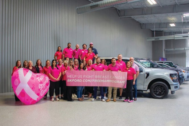 Group in pink shirts holding a breast cancer awareness banner in a garage with cars and a large pink ribbon