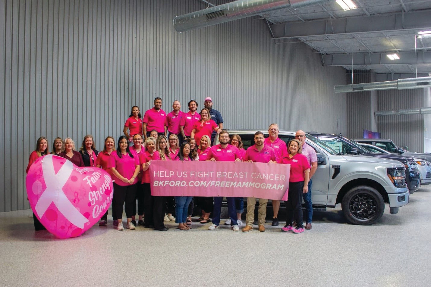 Group in pink shirts holding a breast cancer awareness banner in a garage with cars and a large pink ribbon