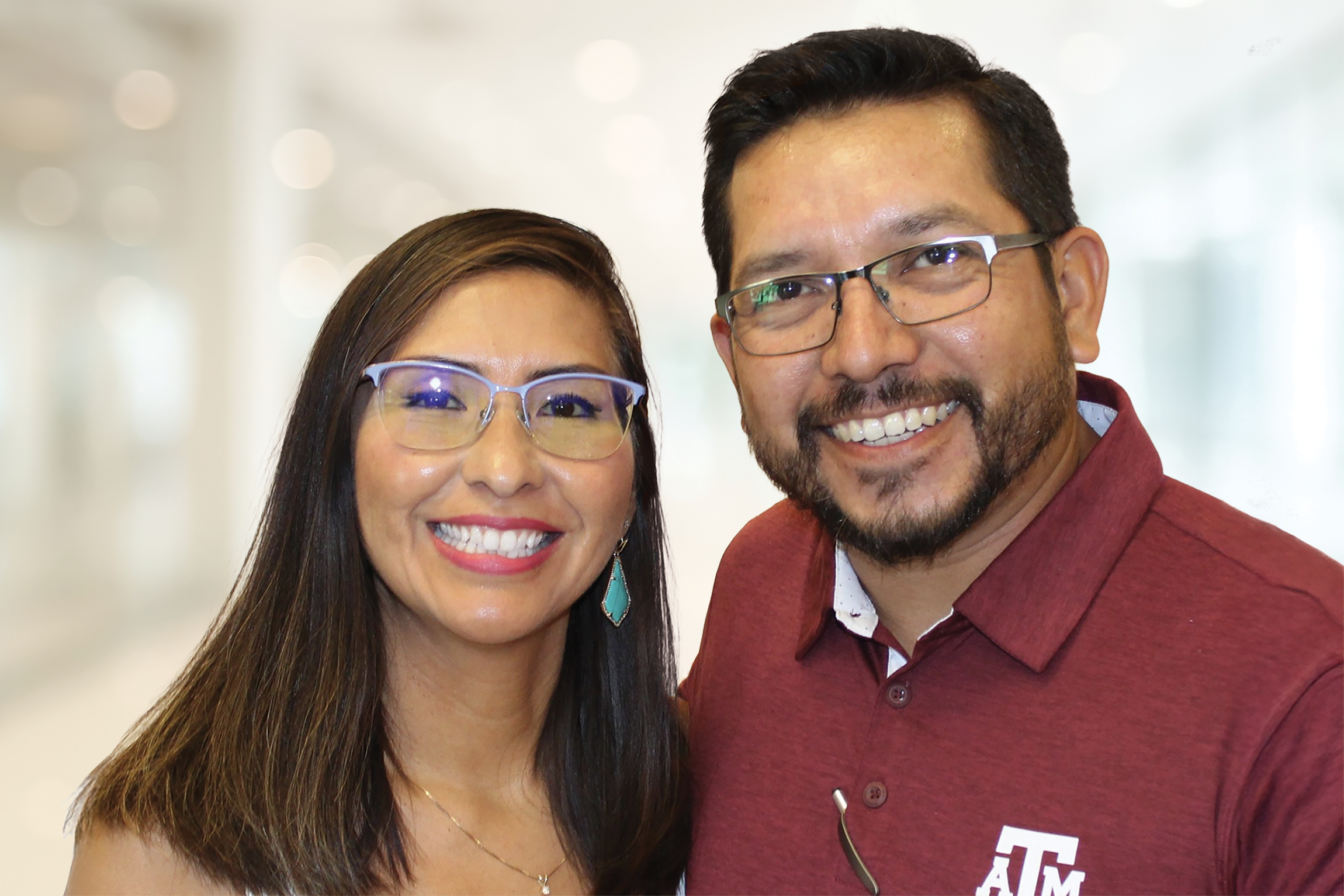 Two people smiling at the camera, one with long hair and glasses, the other with short hair and glasses wearing a maroon A&M shirt, standing in front of a blurred indoor background.