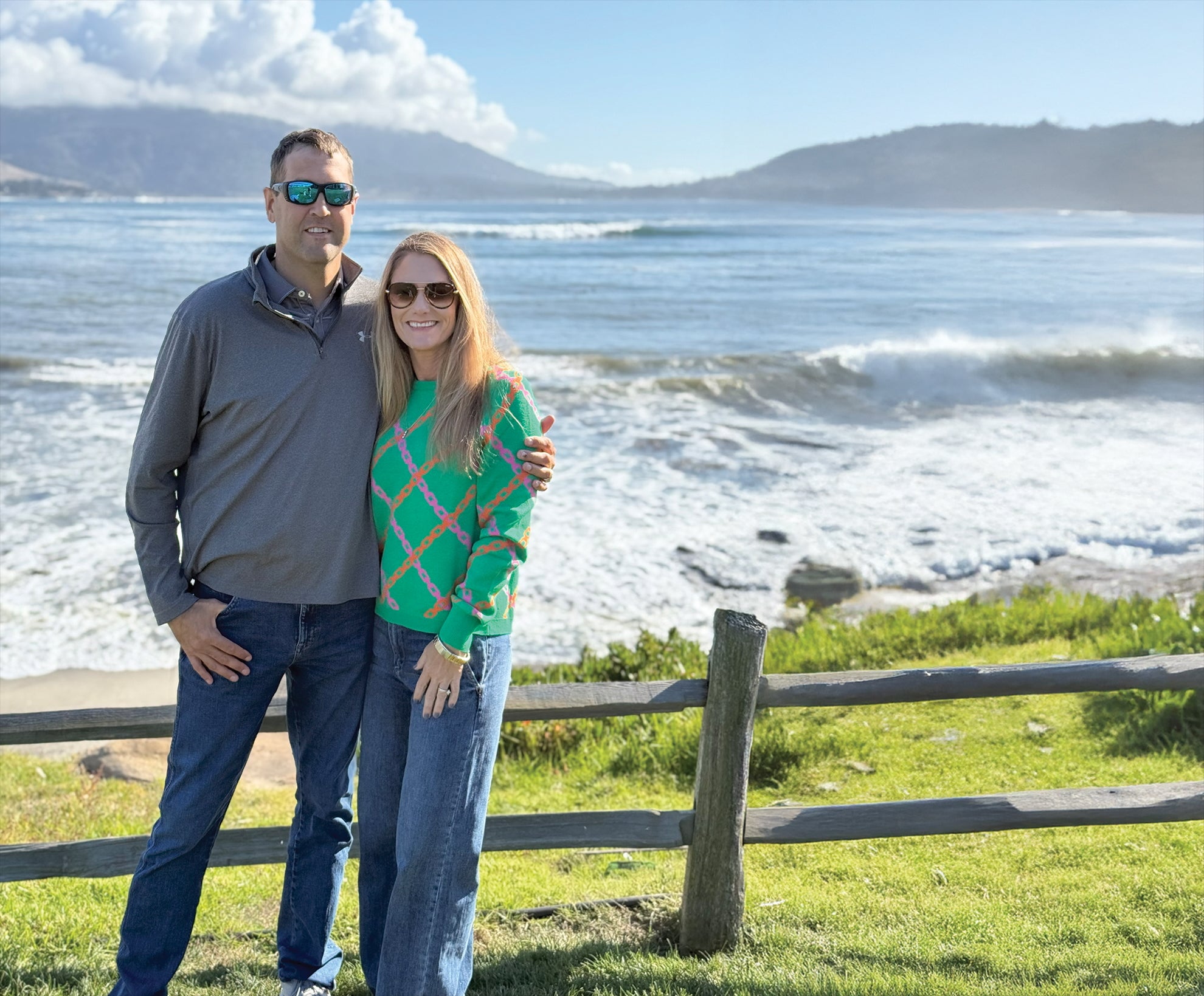Two people stand in front of a wooden fence with ocean waves and mountains in the background on a sunny day.