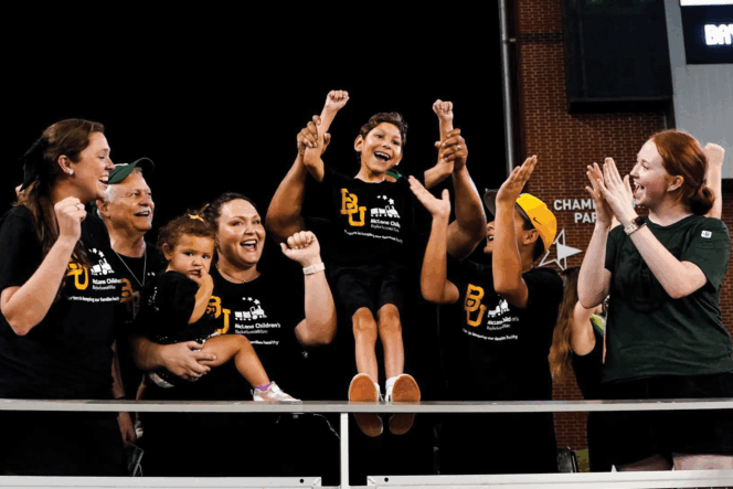Group in matching Baylor Scott & White McLane Children's shirts cheering at Baylor University's McLane Stadium. One adult lifts a disabled child.