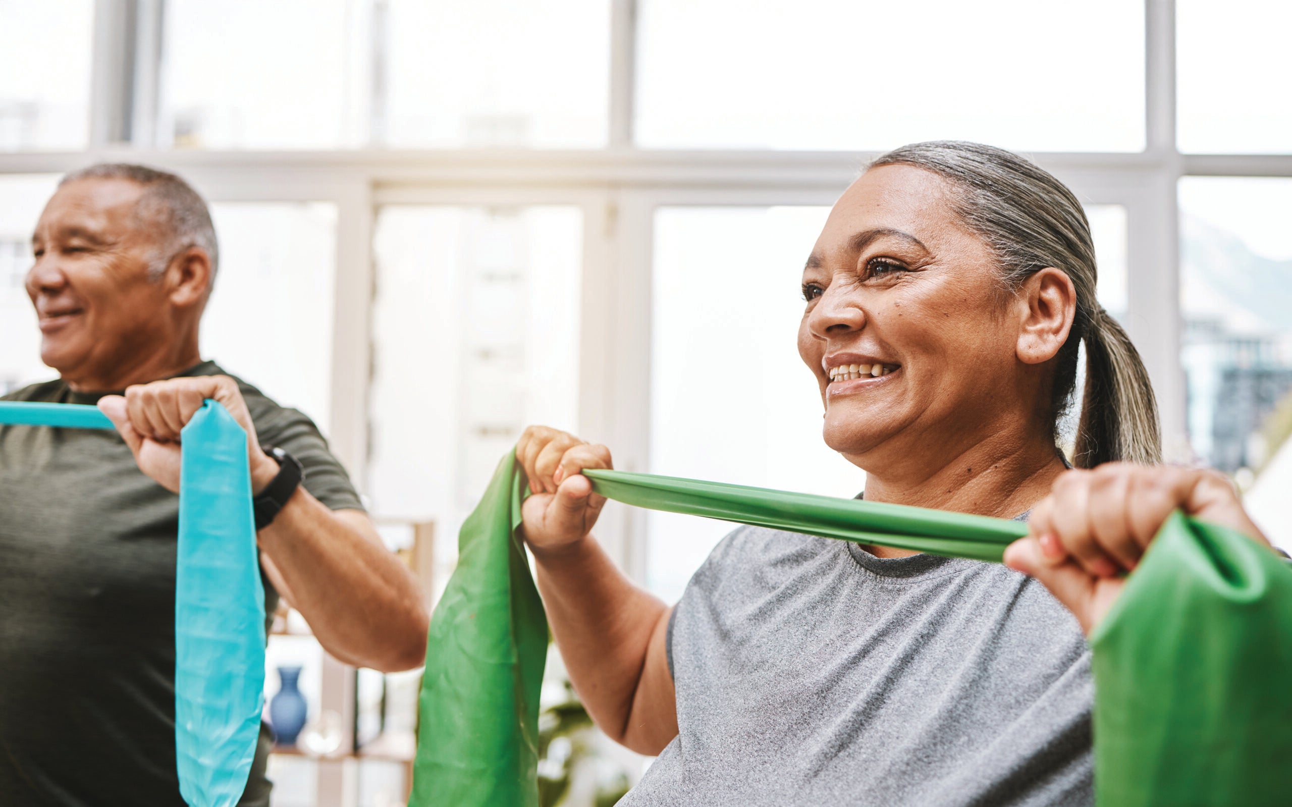 Two adults participate in a wellness class, stretching resistance bands during a guided strength and mobility exercise inside a bright studio.