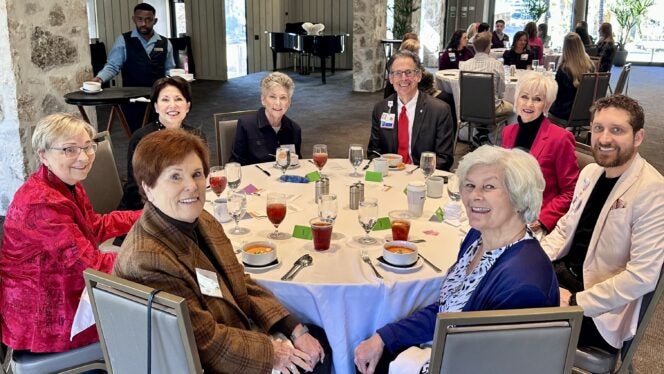 People seated around a round table at a formal luncheon, with place settings and drinks in a bright event space.
