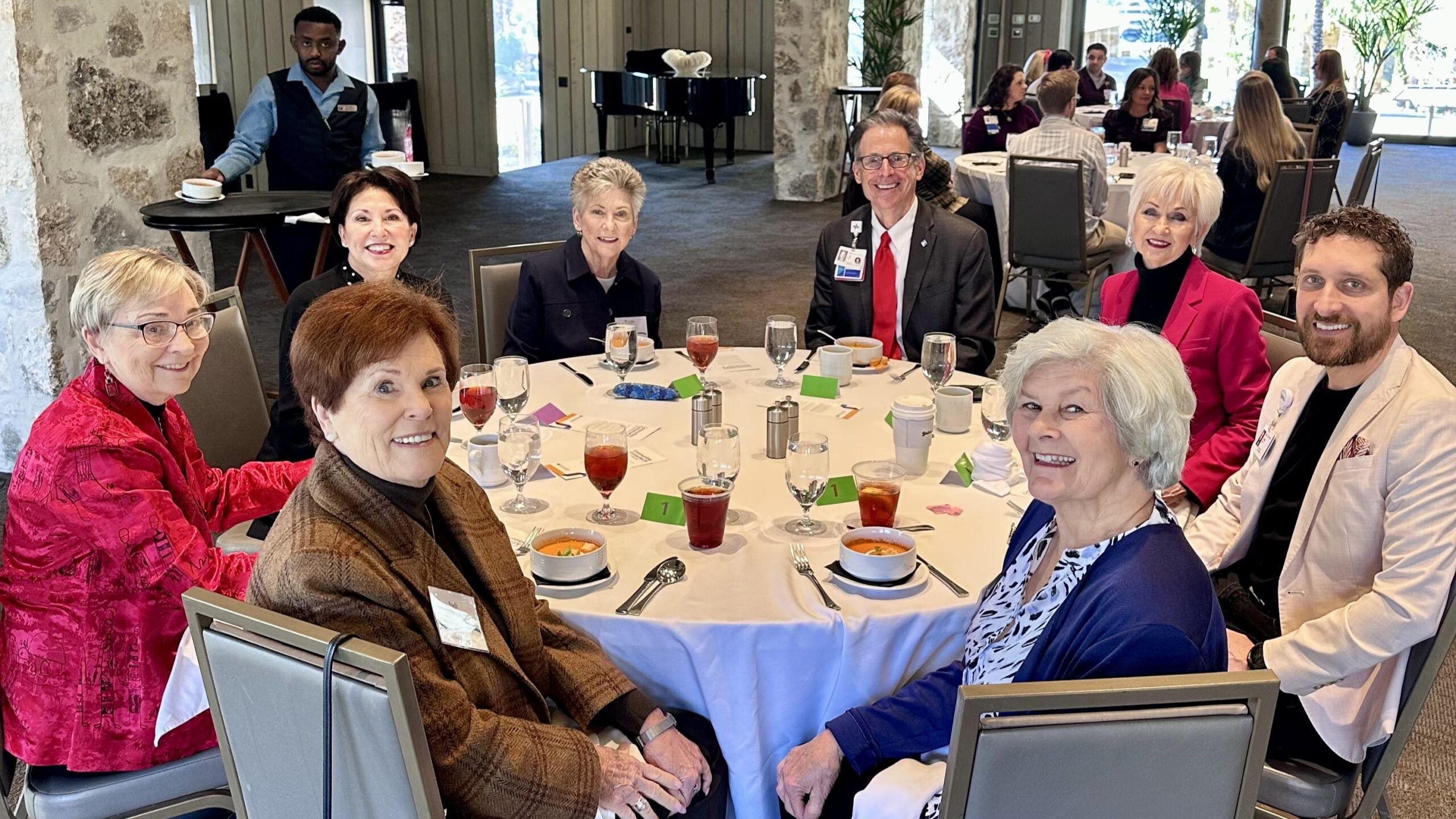 People seated around a round table at a formal luncheon, with place settings and drinks in a bright event space.