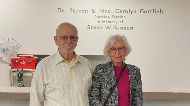 Two people stand at a hospital nursing station with a wall dedication reading “Dr. Steven & Mrs. Carolyn Gottlieb Nursing Station, in memory of Steve Wilkinson.”