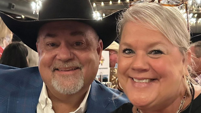Two people seated together at an indoor event under string lights, one wearing a cowboy hat.