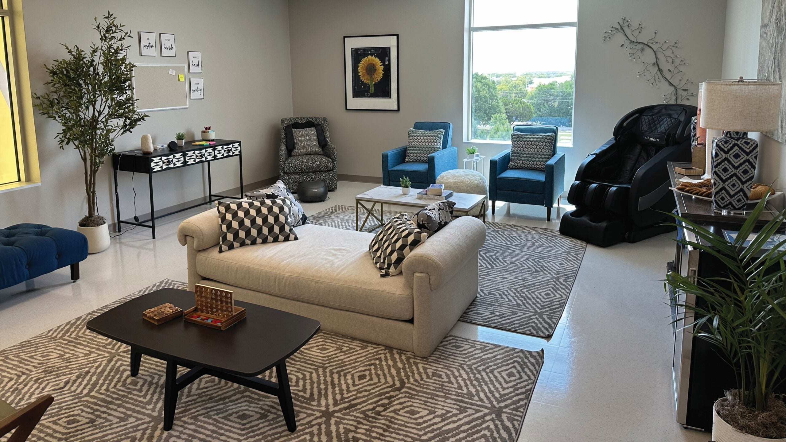 A bright, modern lounge with sofas, armchairs, a massage chair, patterned rugs and soft lighting, designed as a calm, welcoming space.